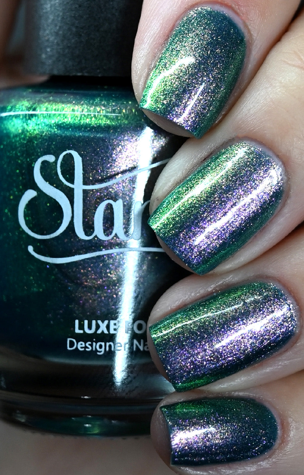 A close-up image of fingernails painted with a green nail polish that has a strong pink, blue, green color-shifting shimmer. The polish is glossy and the brand name is visible on the bottle.